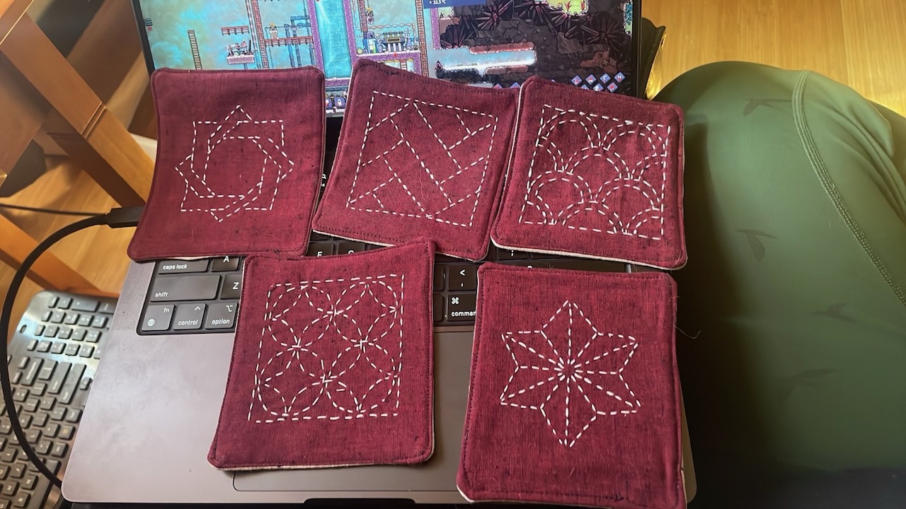 An array of five fabric coasters laid out over a laptop. Each one has white stitching in a traditional sashiko motif. one is interlaced squares, one is staggered rows of arches, one is a 6 pointed star, one is a diagonal nested square pattern, and the last is interlocked rings.