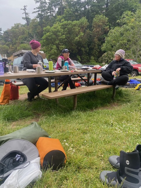 Three women sit surrounded by gear, on a picnic table. The paddle coats and pants are gone, and everyone has warm fleece and hats on. Gear can be strewn in the background.