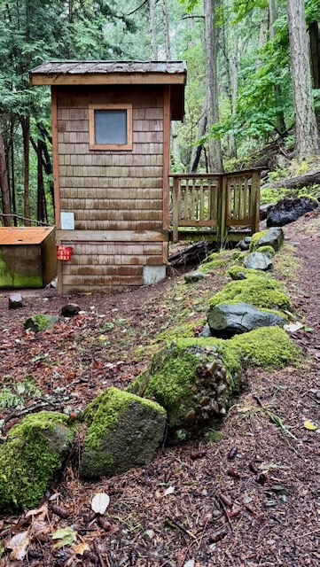 a small wooden shingled structure just large enough for 1 room, with a single frosted window. A small deck allows for entry above ground level. Hung at it's base is a red pail with stenciled text on it. To the right, mossy rocks edge a path up to the deck and entry.