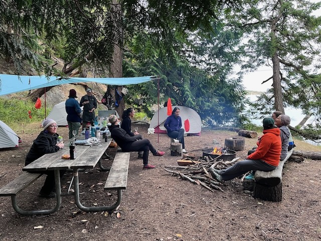 a campsite with a pair of wooden picnic tables, a firepit, and a log to sit on. The area is strewn with women in cold weather gear, and the two guides can be seen in the back, at a camp stove under a tarp strung between trees. There is a fire in the firepit, and the water of Reid Harbor can be seen in the background.