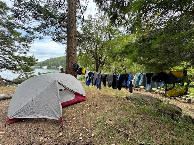a small grey and red two person tent is pitched next to a tree. a clothes line is hung from the tree out of frame, and there are multiple people's paddling clothes and life vests hung from it.