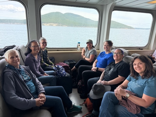 A group of seven women sit in a bank of seats 4 on each side, grinning. behind them out the window is calm water and a green island.
