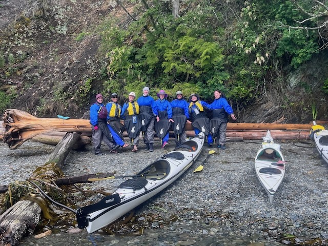 All eight of us are stood in a line in front of a large log of driftwood, behind a kayak. We're all wearing the blue waterproof gear, and our spray skirts which look like black tutus sticking out from around out waists down to our shins. we're grinning and attempting to do a can-can dance.