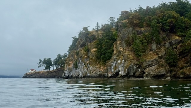 A large cliff with a small lower outcropping of rock. Trees grow along the top of the cliff, and on the outcropping is a very small 1 story white building with a red roof. A small light stands right outside it.
