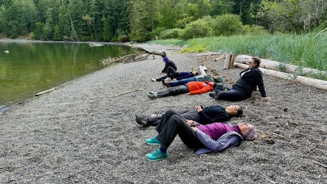 seven women are laid out in a row along a rocky stone beach. the water to the left looks green with the reflection of trees, and to the right is some driftwood and some long mint-green grasses. Everyone looks quite tired.