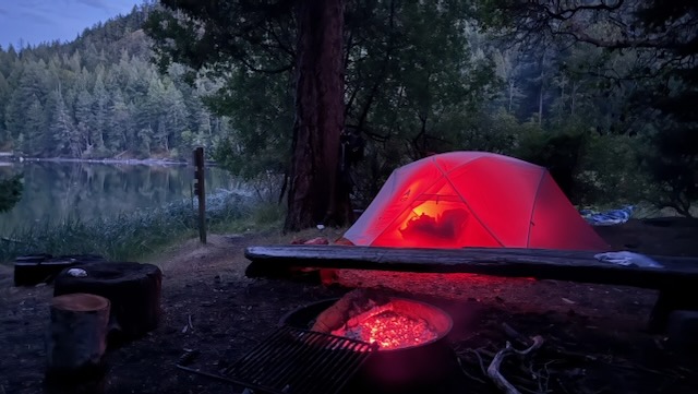 The small tent from the earlier picture, now occupied by it's residents who have a red light on inside. Nearby the coals of the campfire are also glowing red in the dusk light.