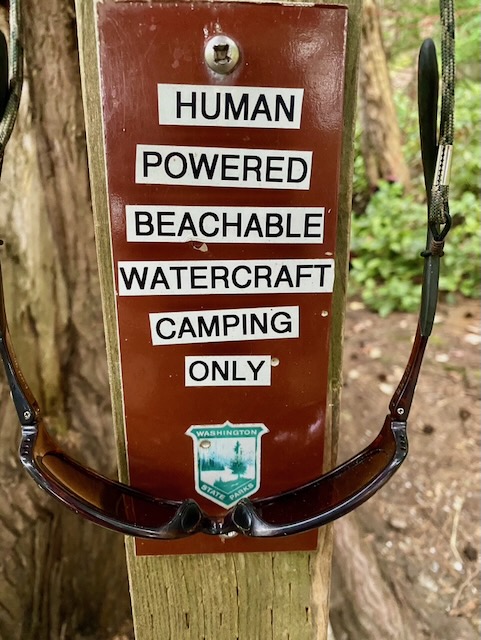 A state park style post and sign, with label maker printed text, reading 'HUMAN POWERED BEACHABLE WATERCRAFT CAMPING ONLY', followed by the Washington state parks logo. Hung over it is a pair of sunglasses.