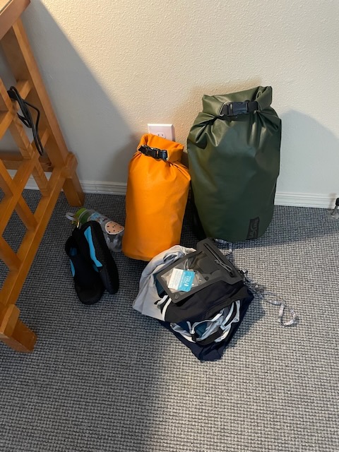 a corner next to a piece of furniture. Stacked against the wall are two full drybags, a smaller orange and a larger green one. Next to it is a stack of clothing for the next day, a waterproof case for a phone, and a water bottle with water shoes clipped to it.