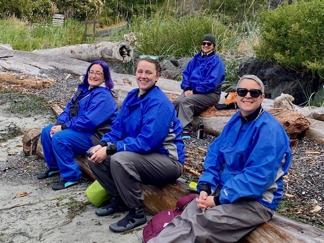 Four women sit on driftwood on a beach, all wearing bright blue paddle coats and pants.