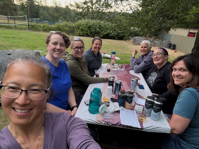 The same women as before are now sitting at a picnic table, which is strewn with water bottles, beer cans, and dice.