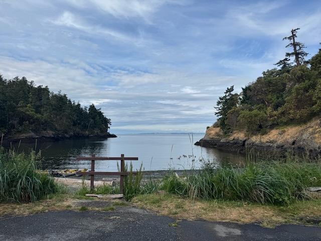 A small inlet, with the Salish Sea beyond. a row of kayaks can be seen on the near edge of the water, and along each side are rocky promontories covered in trees.