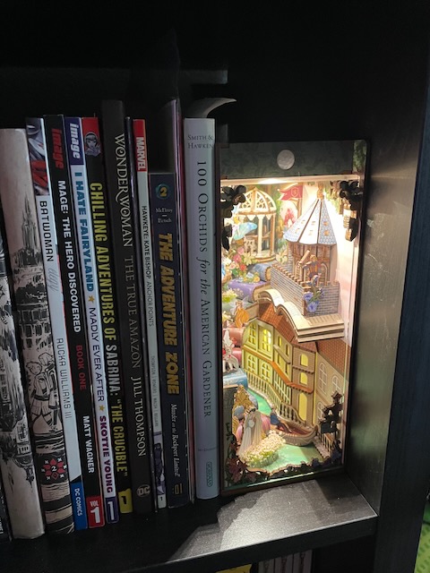 a book shelf with a bunch of books, mostly comic books along with a guide on orchids, and at the end is a small Shakespeare-themed diorama lit up by LEDs. it's got a venitian canal at the bottom, a balcony for romeo and juliet, fairies everywhere, and a staircase made of books that Shakespeare himself is walking up.