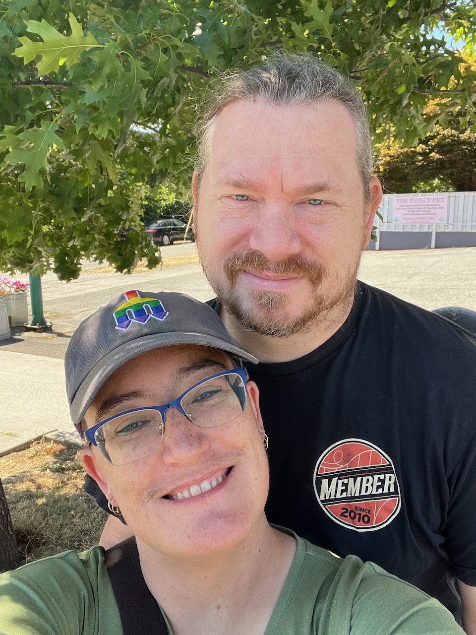 me and my husband taking a selfie, i’ve got a rainbow mariners hat on with blue glasses, he’s rocking the ponytail & scruffy facial hair look, and his chin is as tall as the crown of my head